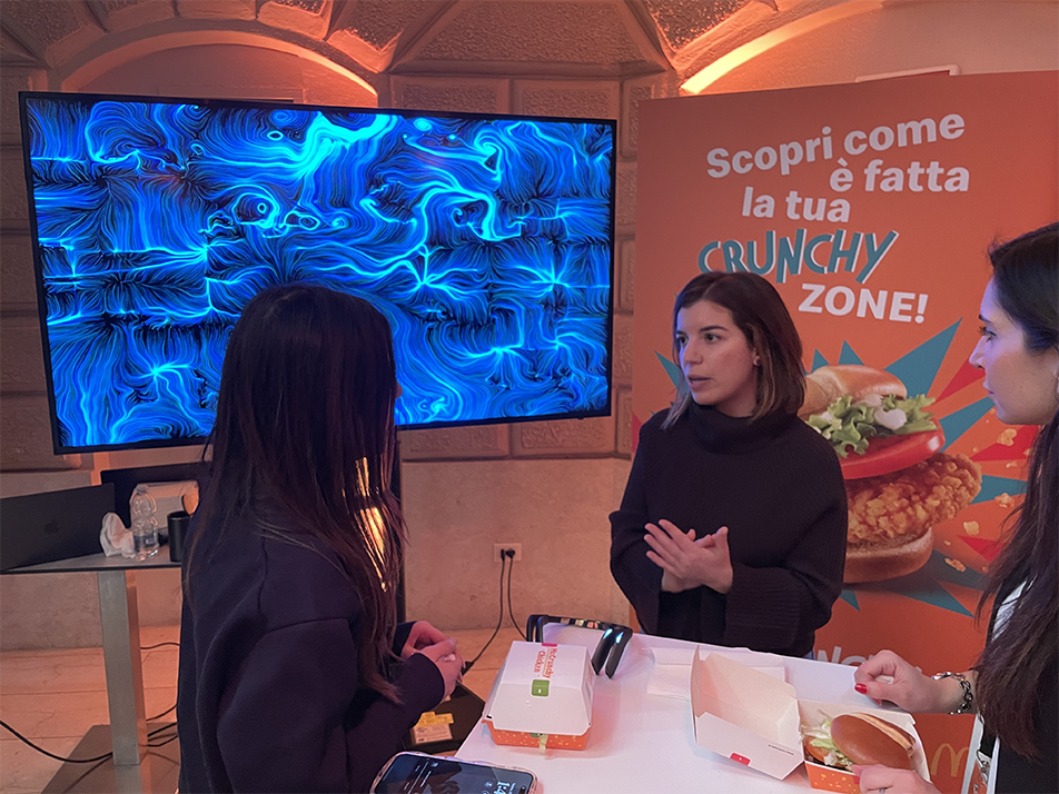 Operatrice BrainArt® spiega l'esperienza neuromarketing a visitatrici durante un evento McDonald's, opera visiva blu sullo schermo